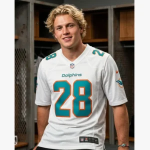 A young man with blonde hair wearing a white De’Von Achane #28 jersey in a locker room setting.