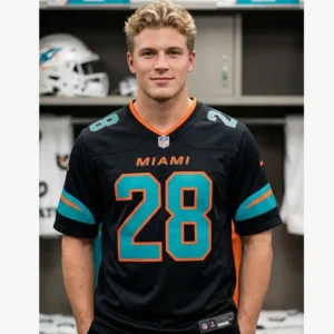 A blonde male model wearing the black De'Von Achane number 28 Dolphins jersey in a locker room setting, showcasing the modern 