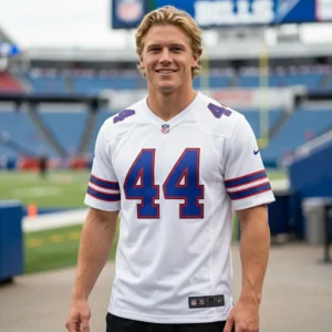 A smiling blonde man standing in a football stadium wearing a white Buffalo Bills jersey with the number 44 in blue and red. The jersey features iconic red and blue striped sleeves and an official NFL patch at the collar.
