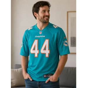 smiling man wearing a turquoise Chop Robinson aqua jersey with white number 44, standing in a modern living room setting with his hands in his pockets.